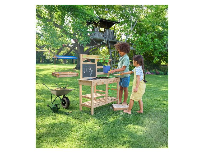Kinderen spelen aan een houten tuintafel met krijtbord, met een boomhut en zandbak op de achtergrond.