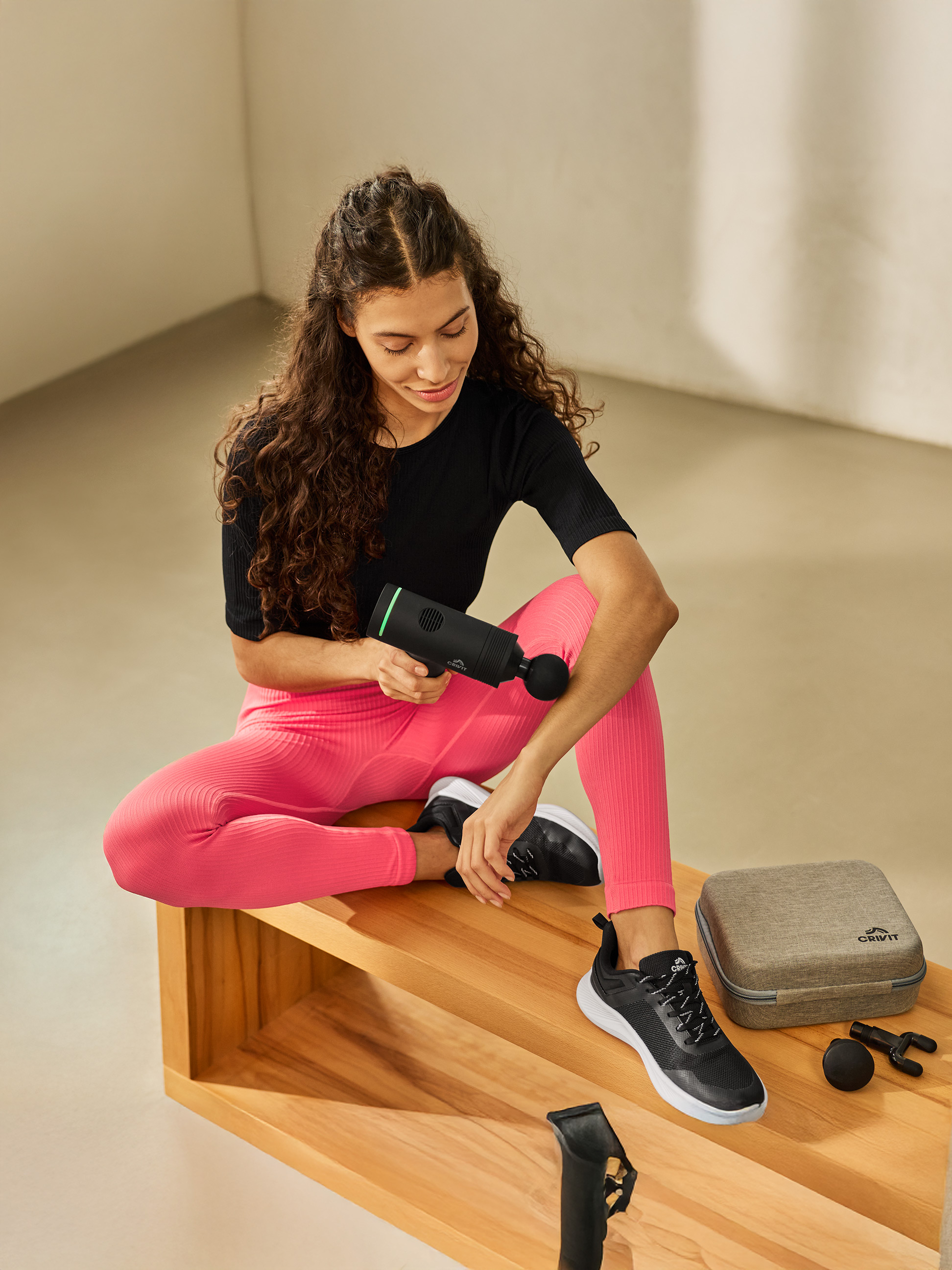 Vrouw gebruikt massagepistool, zittend op houten bank, naast etui en accessoires.