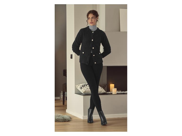 Vrouw in een zwart vest, zwarte legging en zwarte laarzen, staand bij een open haard.