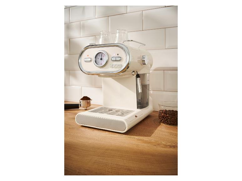 Crèmekleurige espressomachine met manometer en accessoires op een houten aanrecht.