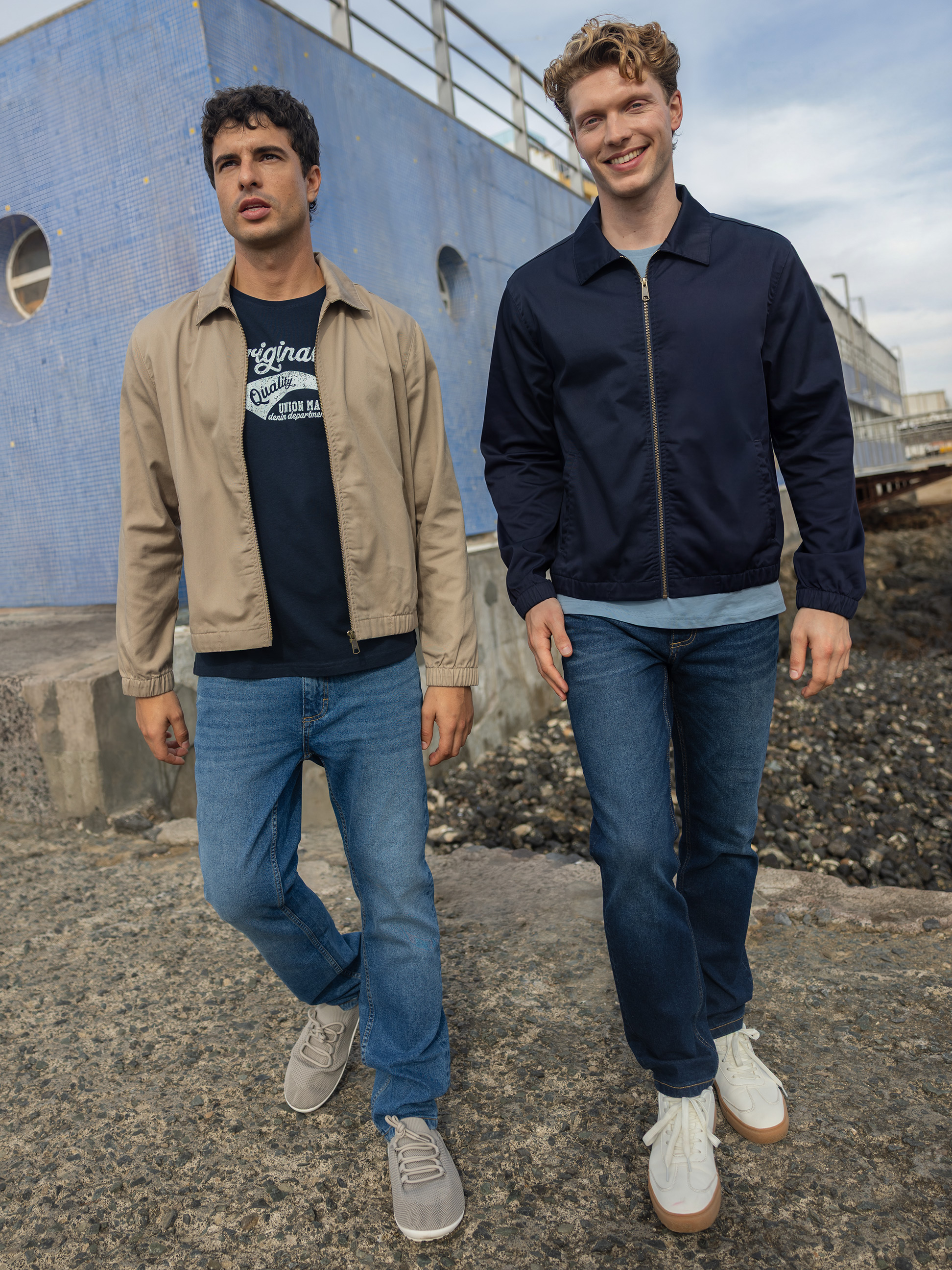 Twee mannen in bomberjacks en jeans lopen langs een blauw gebouw.