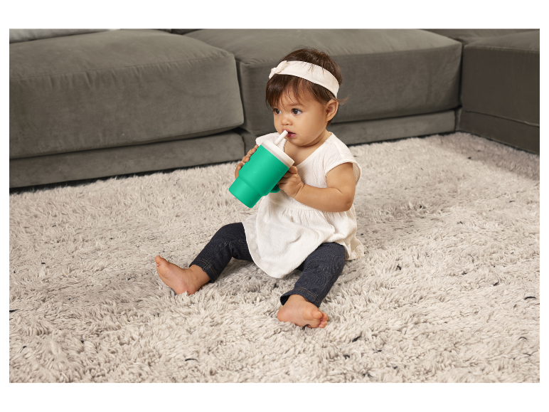 Baby drinkt uit een beker met rietje op een tapijt.