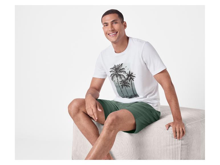 Man in wit T-shirt met palmprint en groene shorts zit op een poef.