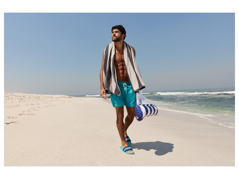 Man in zwembroek en met handdoek loopt op het strand.
