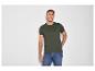 Man in donkergroen T-shirt en blauwe jeans, lachend tegen een witte muur.