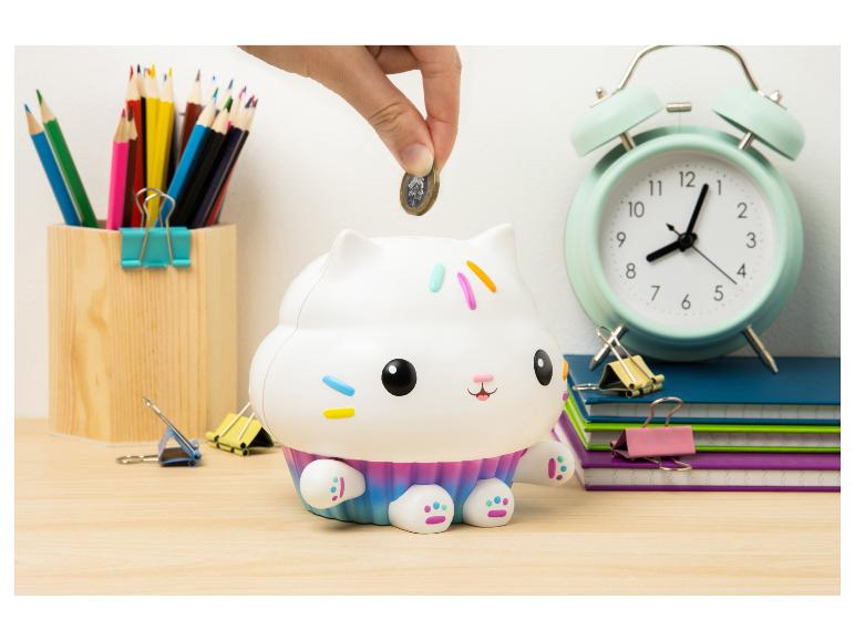 Hand stopt munt in cupcake-kat spaarpot, omringd door schoolspullen.