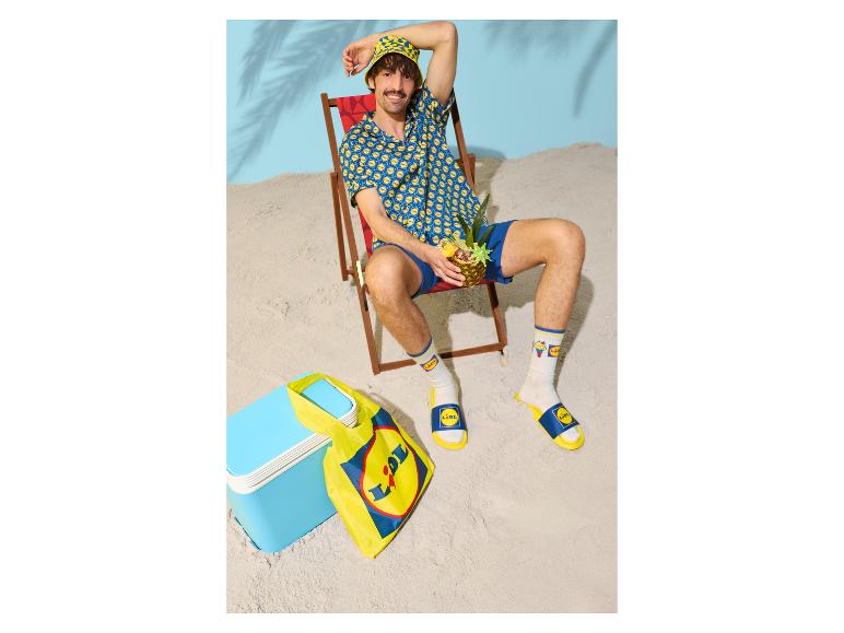 Man in zomerkleding met ananas, naast tas en koelbox op zand.