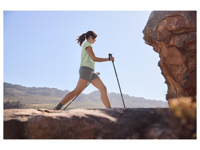 Vrouw in lichtgroen T-shirt en grijze shorts wandelt met trekkingstokken op een rotsachtig pad.