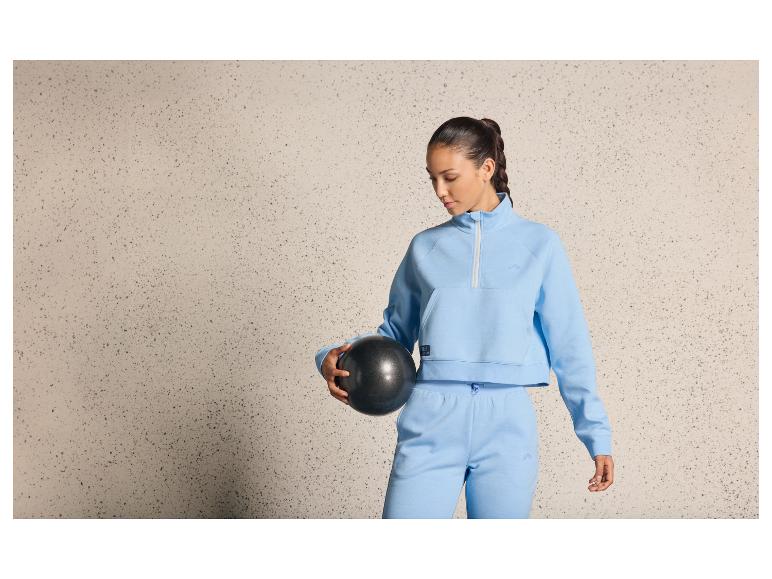 Een vrouw in een blauwe trainingsbroek en een blauwe trui houdt een zwarte bal vast.