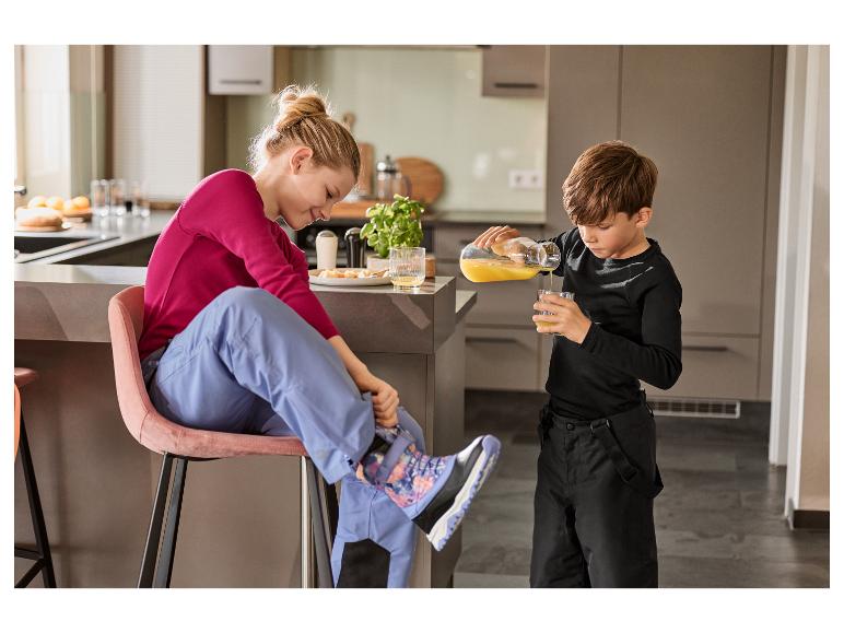 Vrouw en kind in de keuken, kind schenkt sap, vrouw trekt laarzen aan.