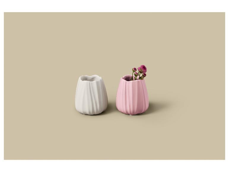 Twee kleine geribbelde vazen, één wit en één roze met bloemen, op een beige achtergrond.