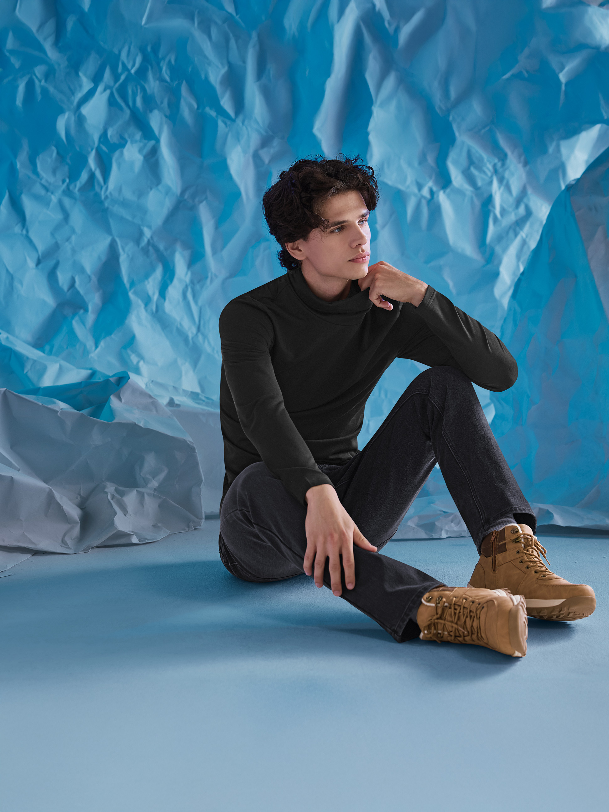 Man in een zwarte coltrui, jeans en bruine laarzen, zittend op een blauwe achtergrond.