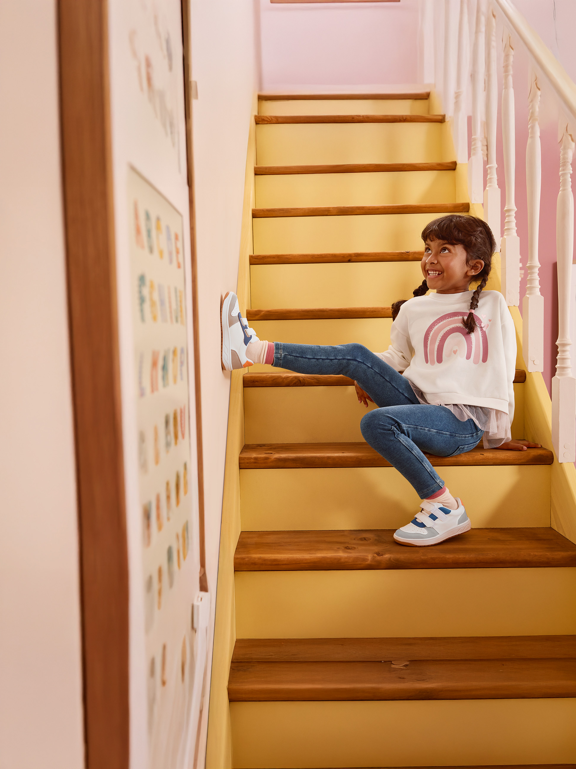 Meisje in witte regenboogtrui en blauwe jeans zit op houten trap