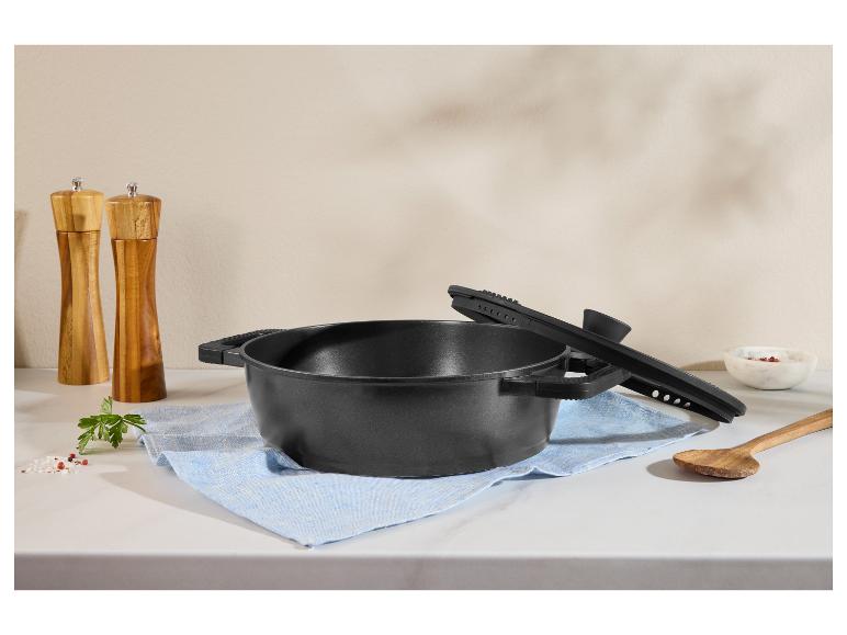 Zwarte pan met deksel, zout- en pepermolens, houten lepel en kruiden op een aanrecht.