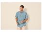 Man in blauw overhemd met korte mouwen en beige korte broek, zonnebril vasthoudend.