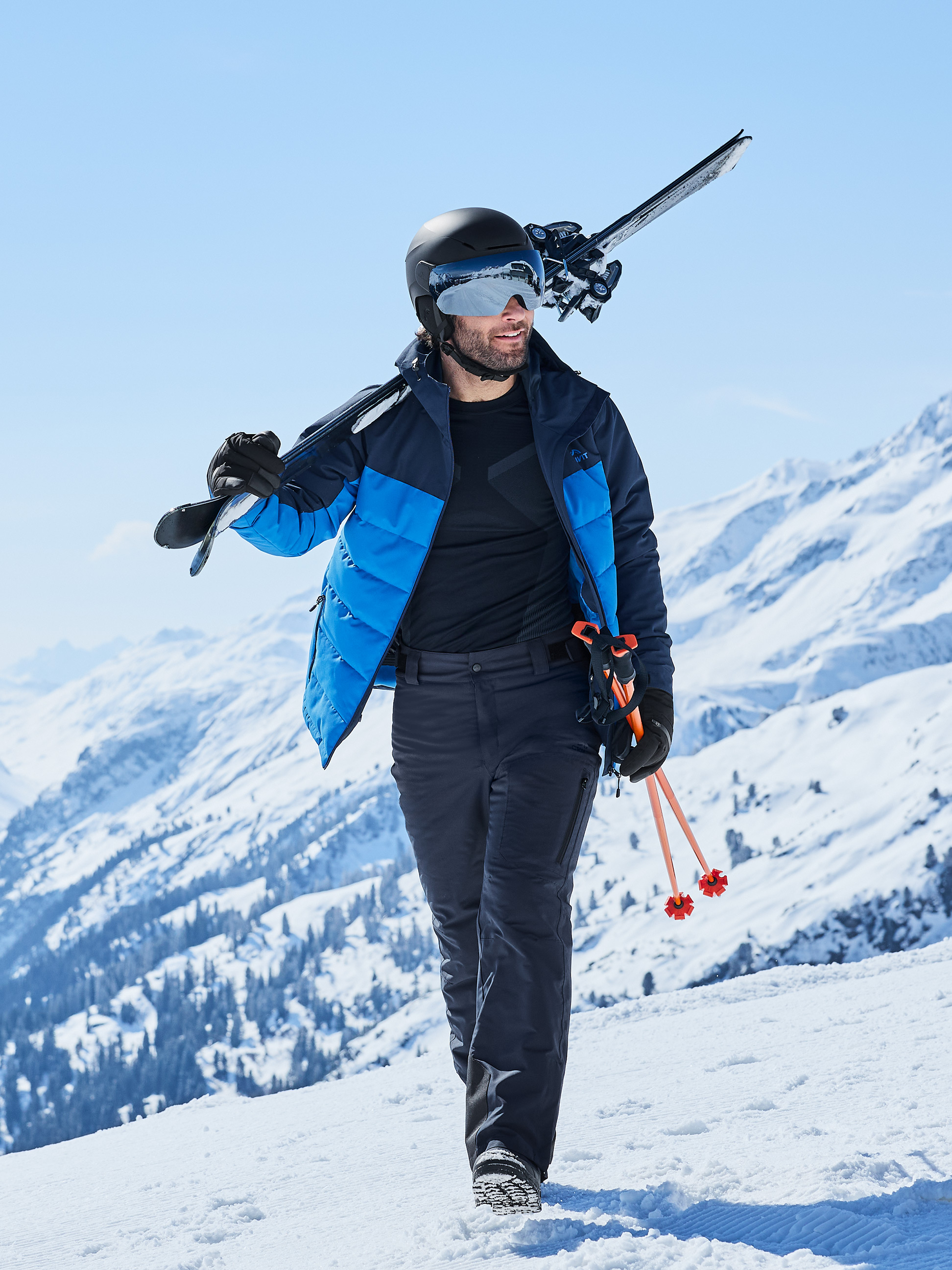 Man in skijas, broek en helm met ski's en stokken in de sneeuw.