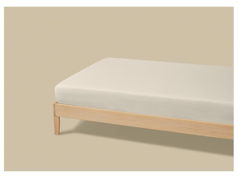 Een lichtgekleurd matras op een houten bedframe tegen een beige achtergrond.