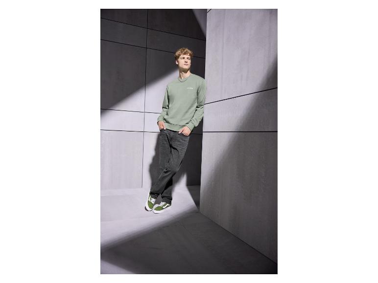 Man in groene sweater met 'Club 1990' en donkere jeans, leunend tegen een muur.