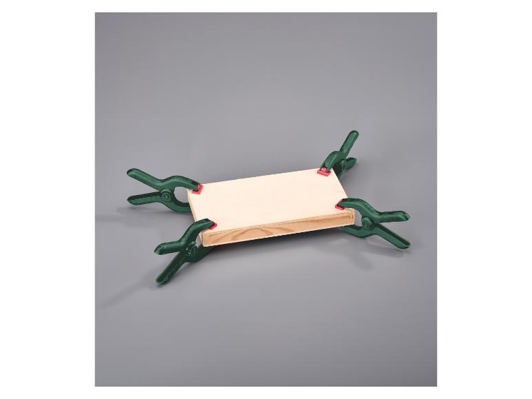 Vier groene veerklemmen bevestigen een houten plank op een grijze achtergrond.