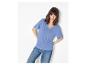 Vrouw in een blauw T-shirt met korte mouwen en jeans, leunend tegen een witte muur.