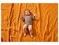 Lachende baby in een bruine romper, liggend op een oranje getextureerde deken.
