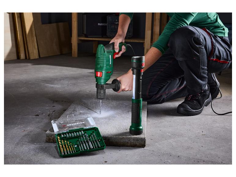 Een persoon gebruikt een groene boormachine op een betonplaat, met een boorset en een zaklamp ernaast.