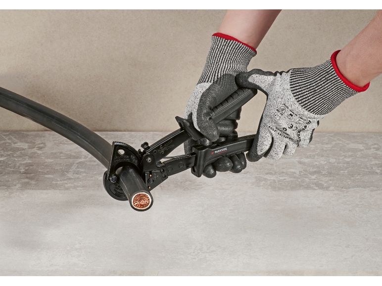 Een persoon snijdt een dikke kabel met een ratelkabelknipper en draagt werkhandschoenen.