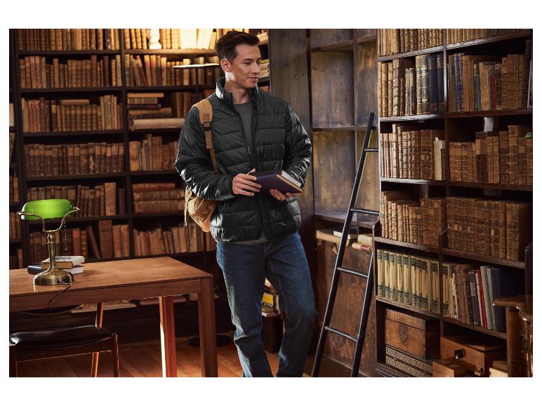 Man in een zwarte pufferjas en jeans, met boeken in een bibliotheek.