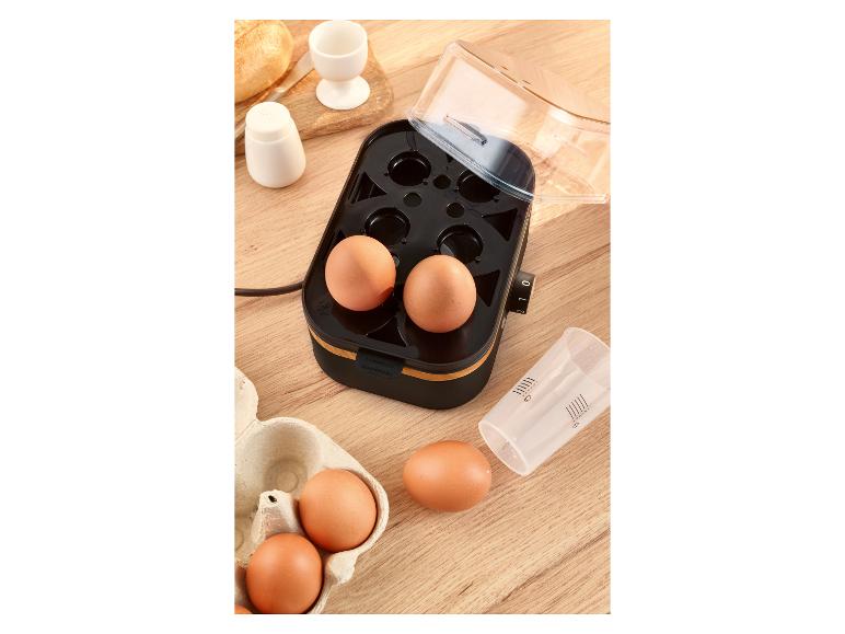 Eierkoker met eieren, maatbeker en brood op een houten tafel.