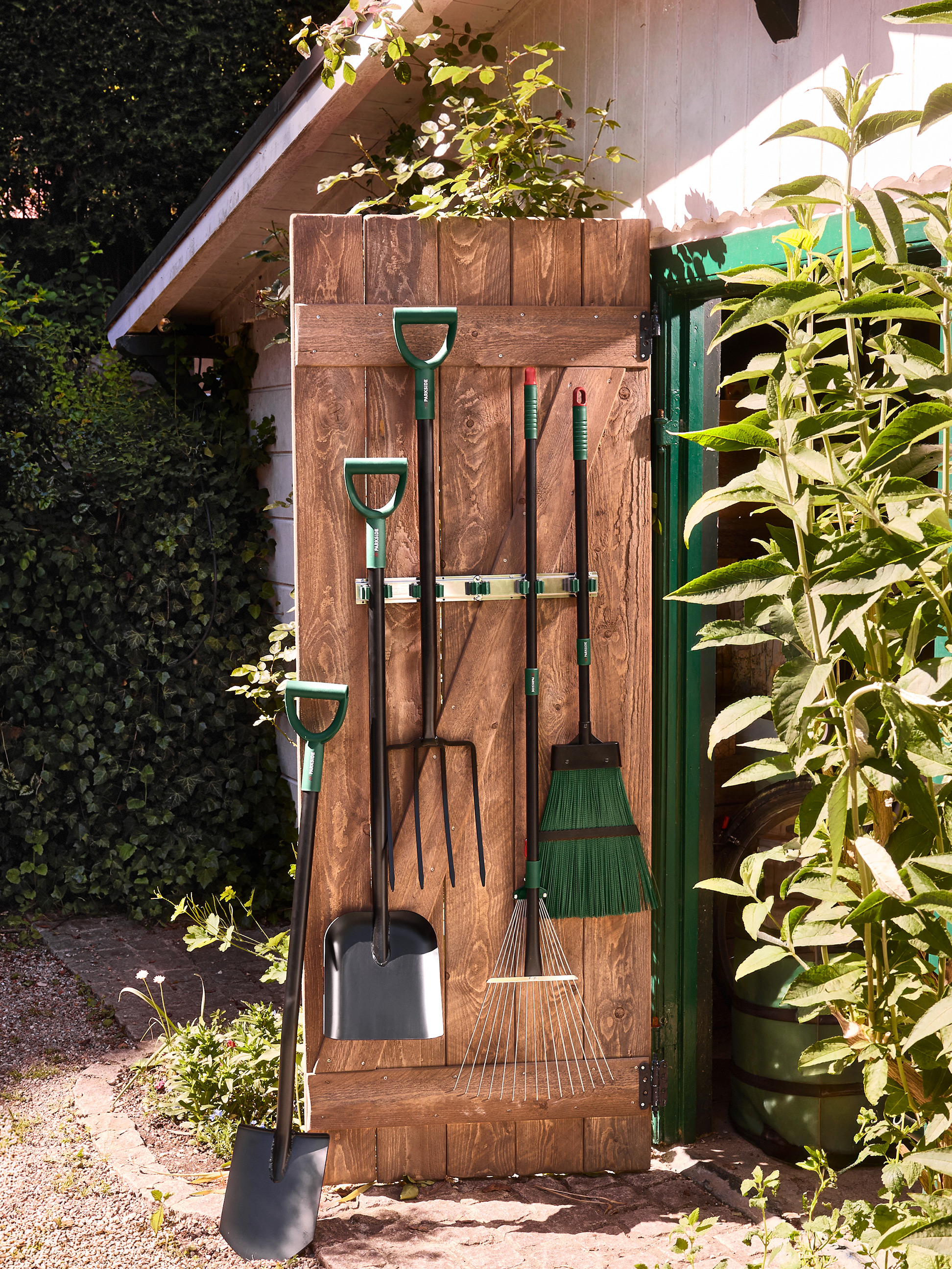 Tuin gereedschap: schoppen, vorken, harken en een bezem, hangend aan een houten schuurdeur.