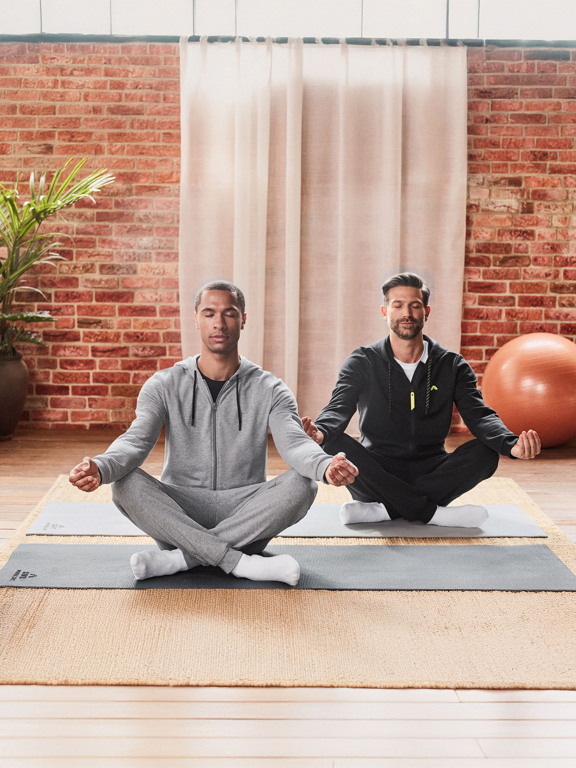 Twee mannen in trainingspakken mediteren op yogamatten in een kamer met een bakstenen muur.