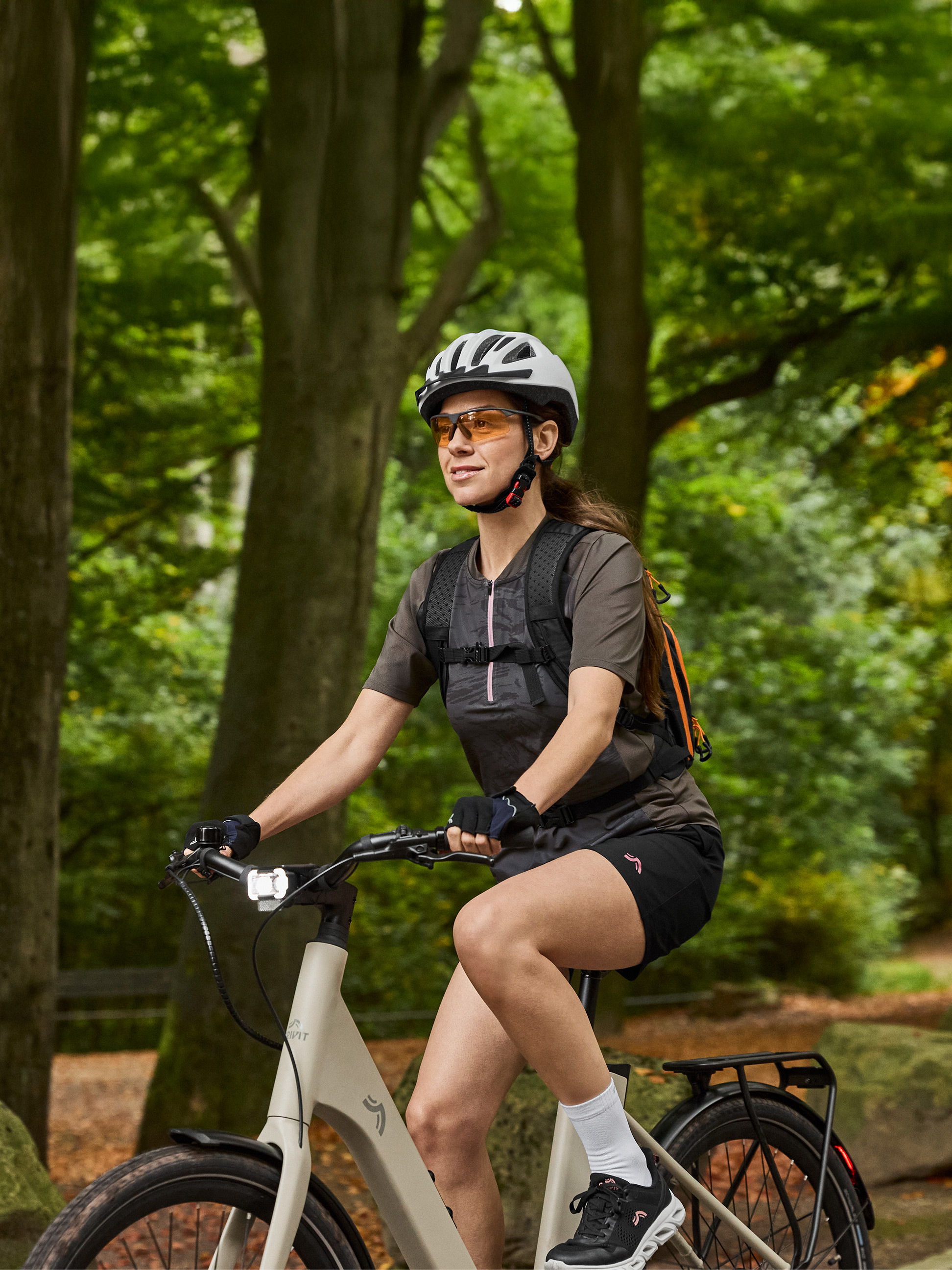 Vrouw op e-bike met helm, zonnebril en rugzak in een bos.