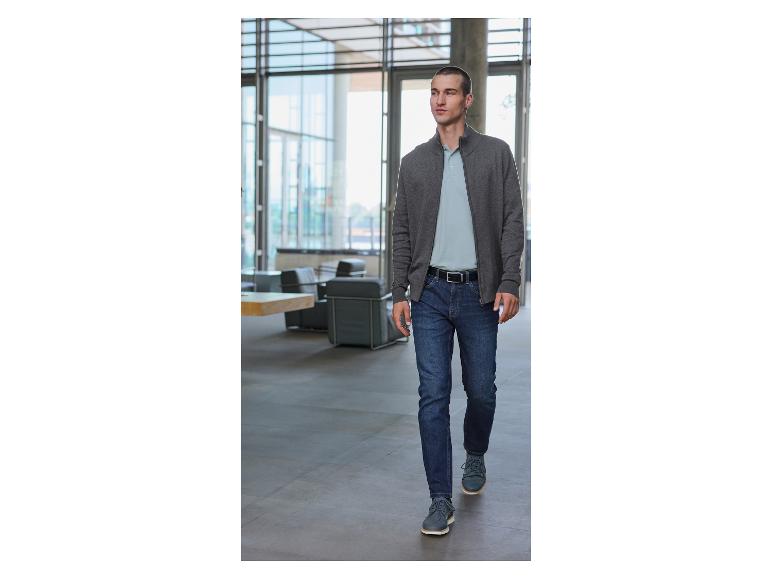 Een man loopt door een modern gebouw, gekleed in een grijze trui met rits, een blauwe jeans en donkergrijze schoenen.