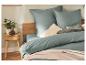 Bed met groen beddengoed, beige sprei en kussens, naast een nachtkastje met plant.