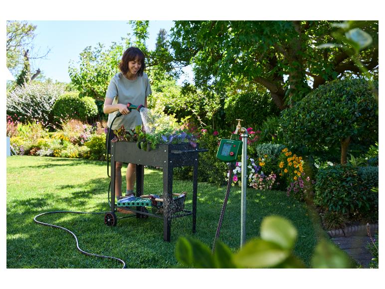 Vrouw besproeit planten in een verhoogde moestuin, naast een besproeiingstimer.