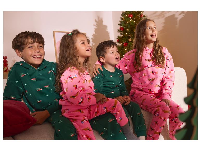 Vier kinderen in feestelijke pyjama's, twee in groen met auto's en twee in roze met teckels, kijkend naar een kerstboom.