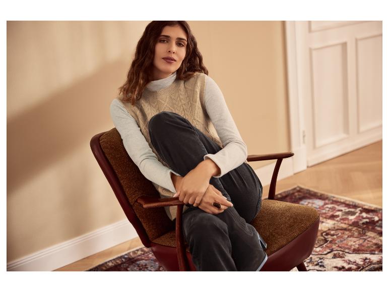 Vrouw in beige gebreid vest en donkere jeans zit in een fauteuil.