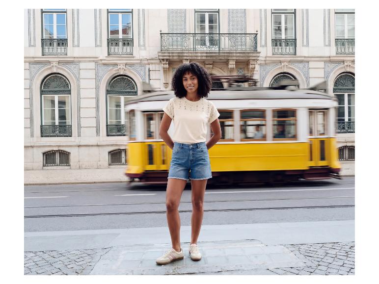 Vrouw in geborduurd T-shirt en denim shorts voor een rijdende gele tram.