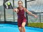 Vrouw in bordeauxrode sportjurk speelt padel met zwart racket en gele bal.