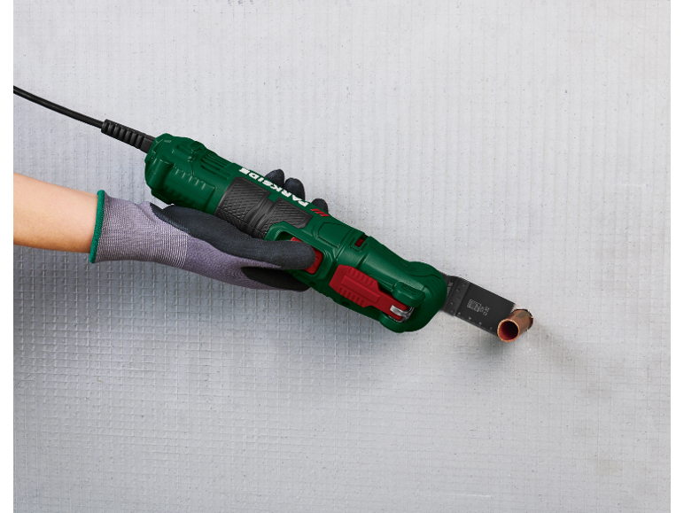 Een hand in een werkhandschoen houdt een multitool vast die een koperen pijp in een muur snijdt.