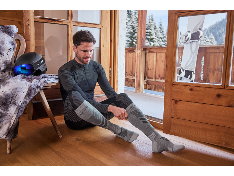 Man in thermisch ondergoed en skisokken, zich voorbereidend op skiën op een besneeuwd balkon.