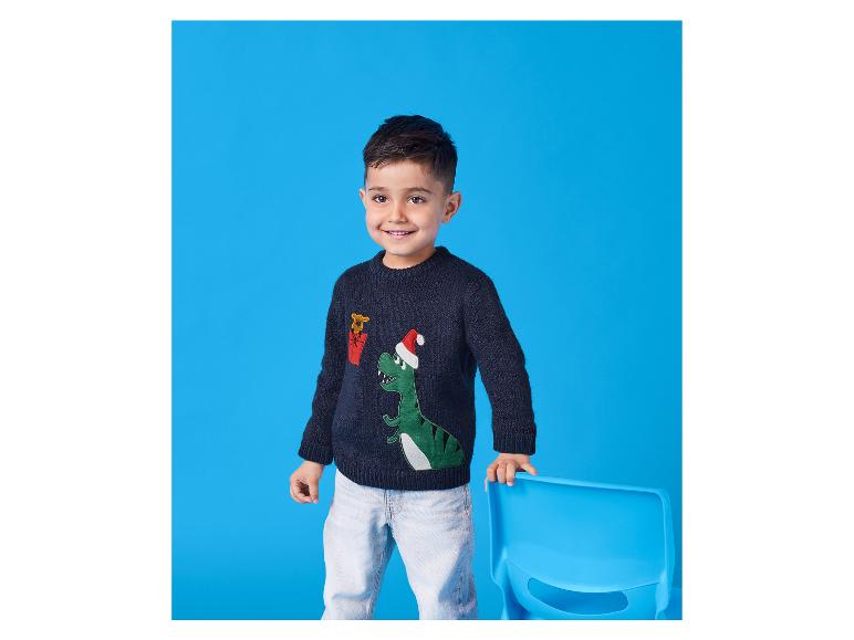 Een lachende jongen draagt een dinosaurus kersttrui en jeans, leunend op een stoel.