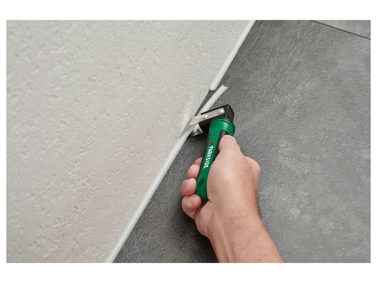 Hand snijdt een witte kitrand tussen een lichte muur en donkere vloer met een groen gereedschap.