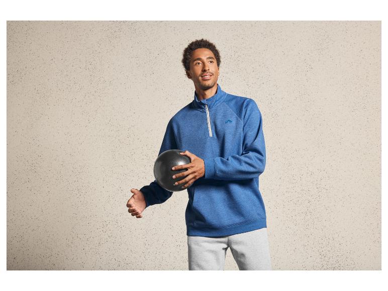 Een man in een blauwe trui met capuchon en grijze joggingbroek, met een zwarte bal in zijn hand.