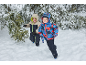 Twee blije kinderen in kleurrijke skipakken rennen door de sneeuw tussen de bomen.