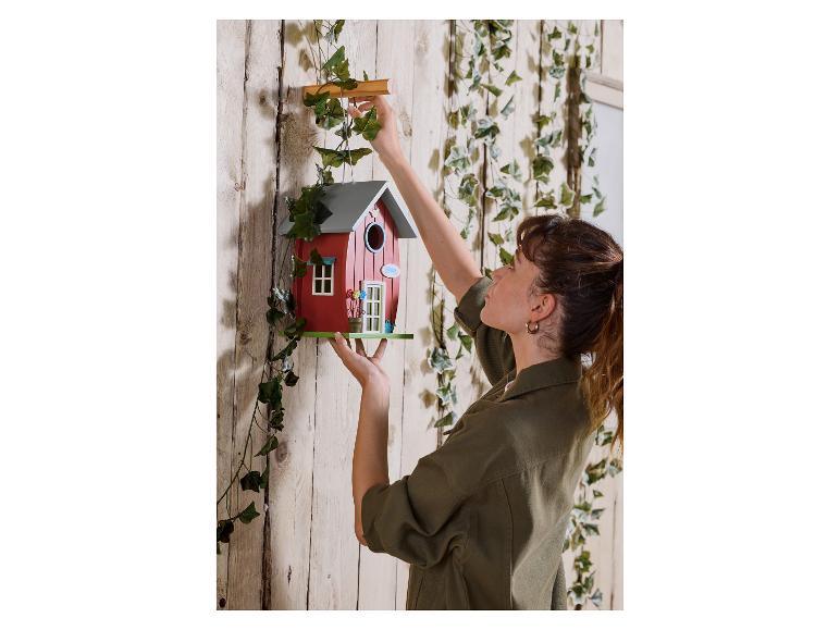 Vrouw hangt een rood vogelhuisje aan een houten muur met klimop.