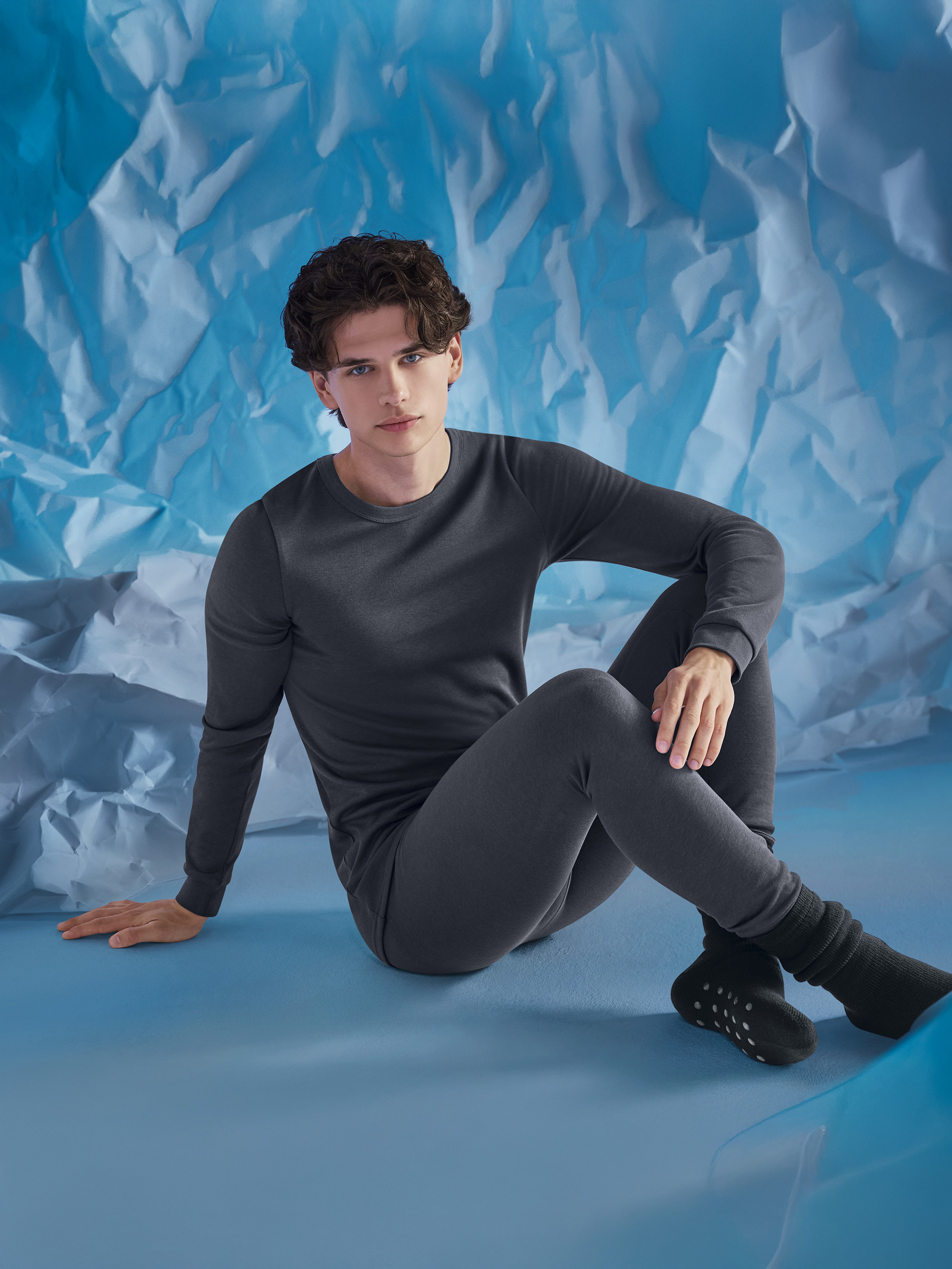 Man in grijs thermisch ondergoed, zittend op een gekreukelde blauwe achtergrond.