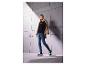 Vrouw in zwarte trui met print, jeans en sneakers, met beige jas over haar schouder.