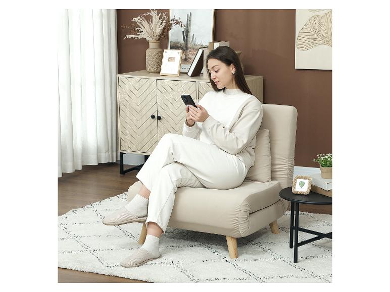 Vrouw zit op beige fauteuil, gebruikt telefoon, in woonkamer met kast en vloerkleed.