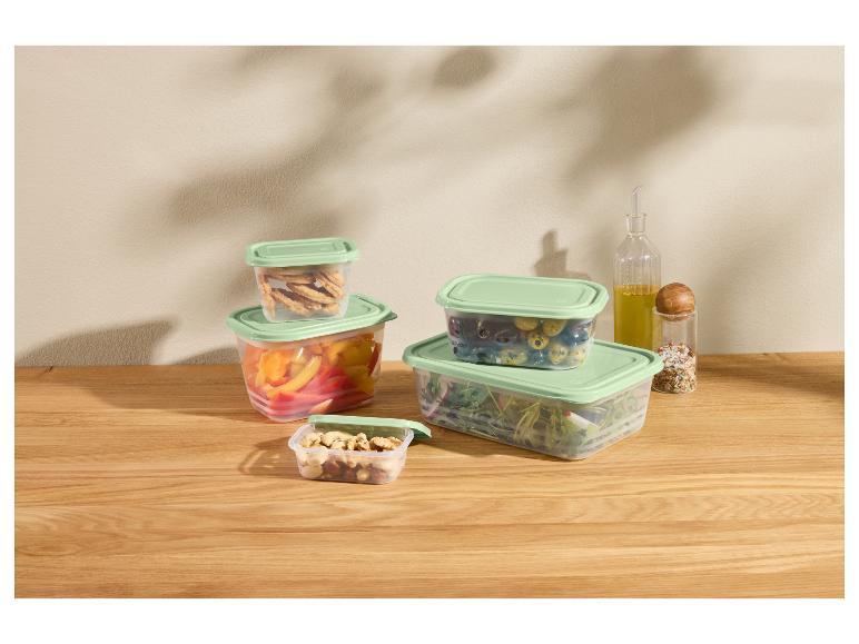 Plastic bakjes met groene deksels gevuld met groenten, noten en crackers op een houten tafel.